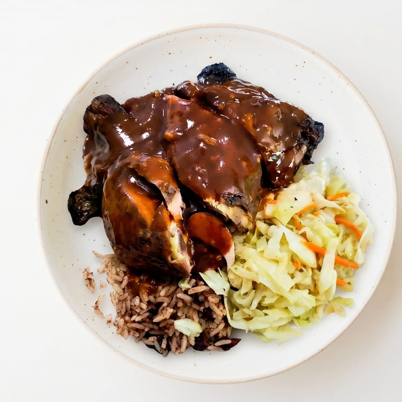 Jerk chicken covered in brown sauce, served with rice and peas, and steamed cabbage.