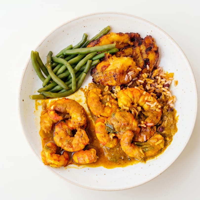 Curried shrimp with rice and beans, green beans, and fried plantains on a white plate.