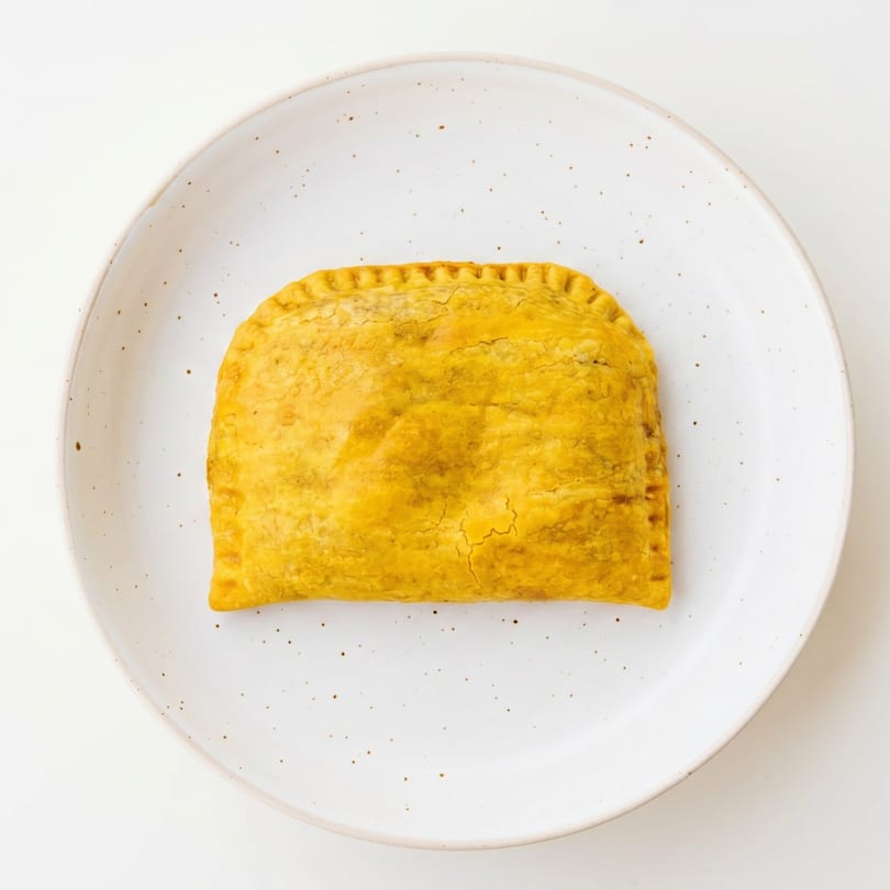 A golden-brown Jamaican beef patty with crimped edges rests on a speckled white plate.