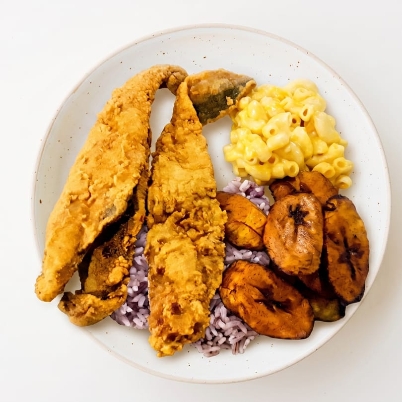 Fried fish, purple rice, macaroni and cheese, and plantains on a white plate.